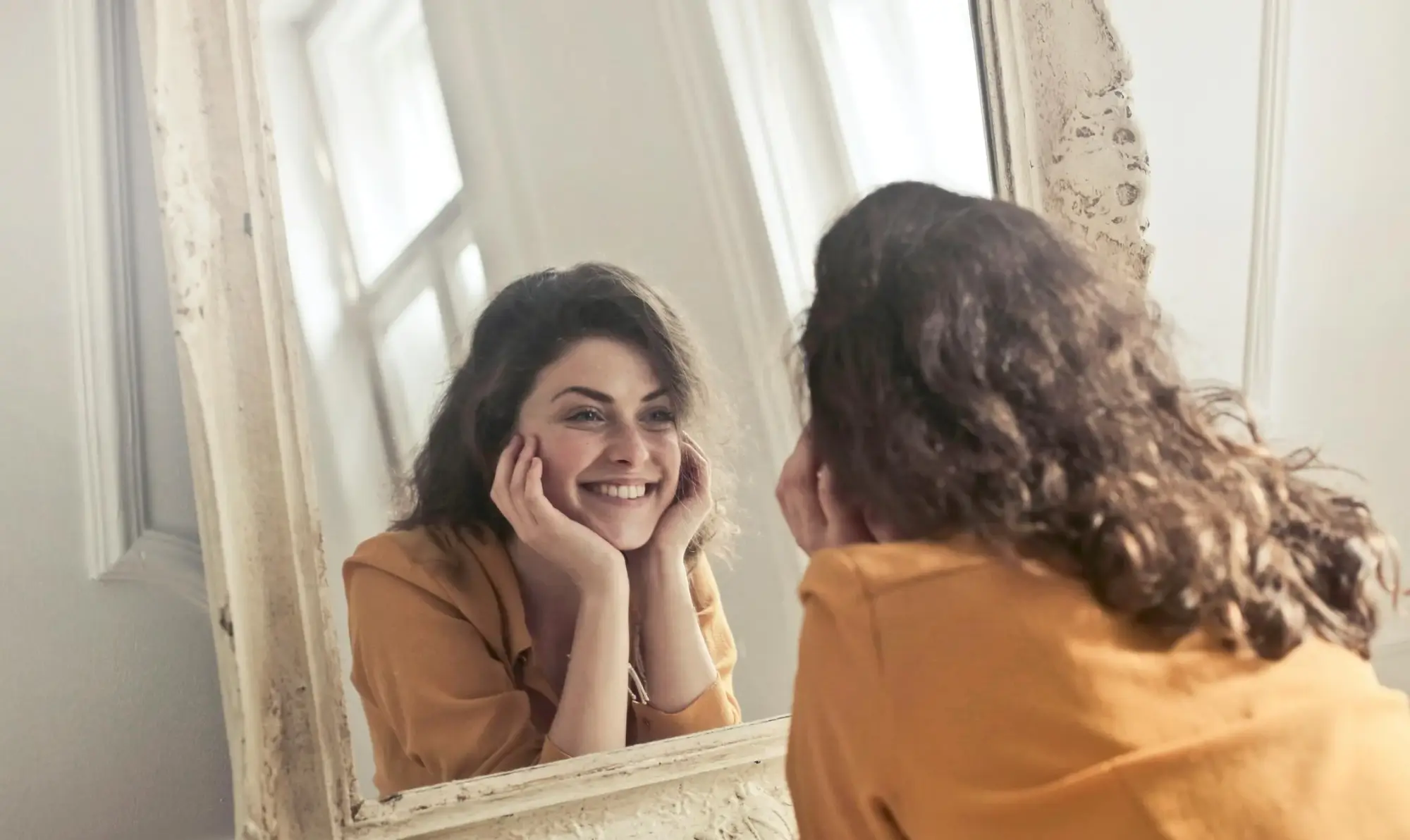 Woman smiling at her reflection in a mirror while holding her face with her hands